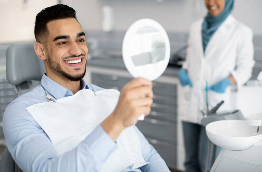 A man sitting in a dentist's chair, holding up a mirror and smiling with his dentist in the background
