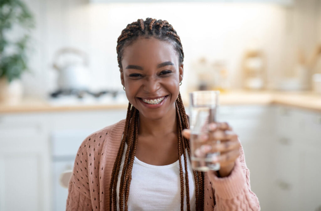 A young woman sitting in a kitchen and smiling with braces as she holds up a glass of water
